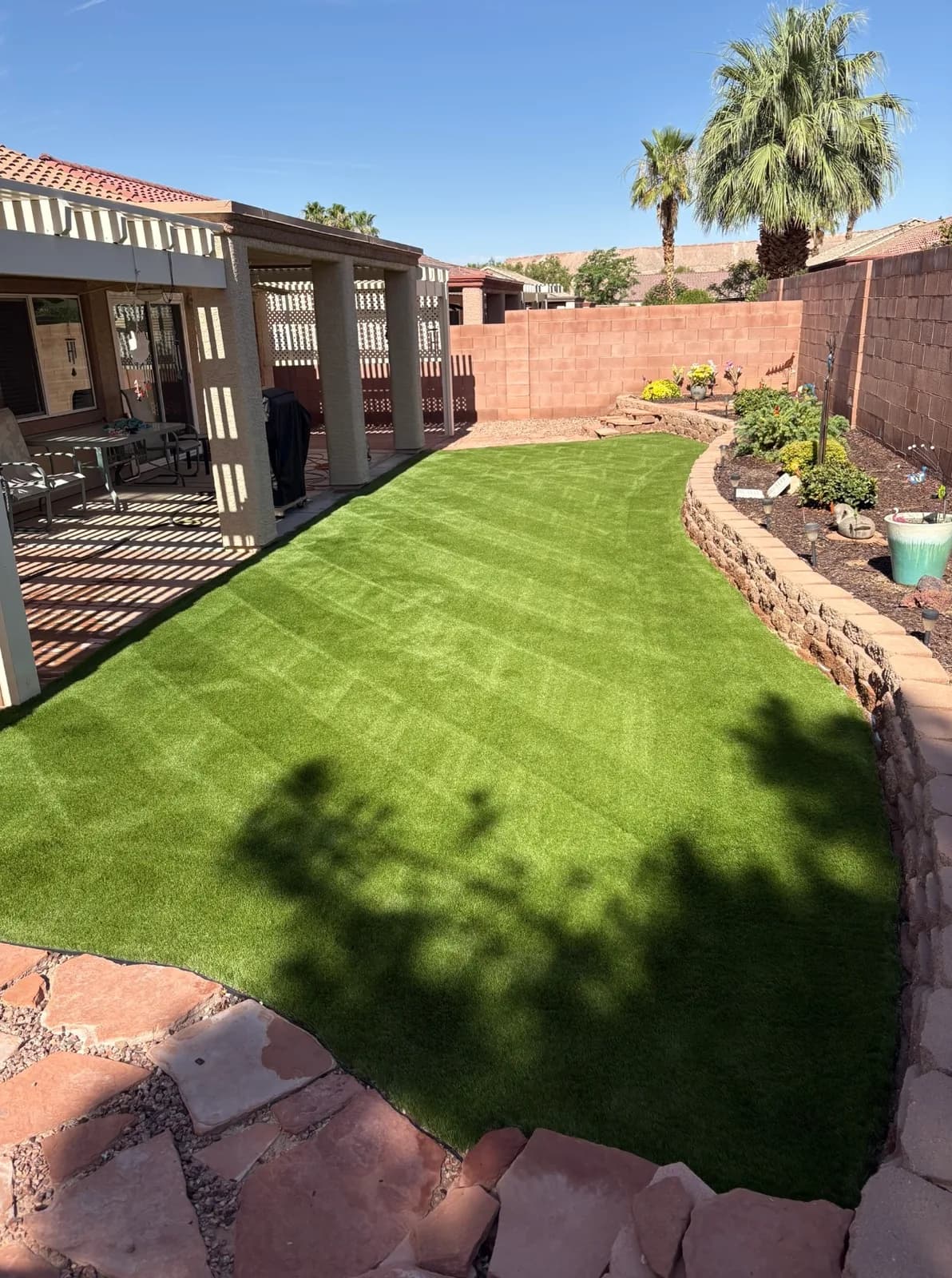Clean artificial turf in a curved Mesquite NV backyard after deep cleaning, bordered by raised stone garden beds, a pergola patio, and block wall fencing with palm trees in background.