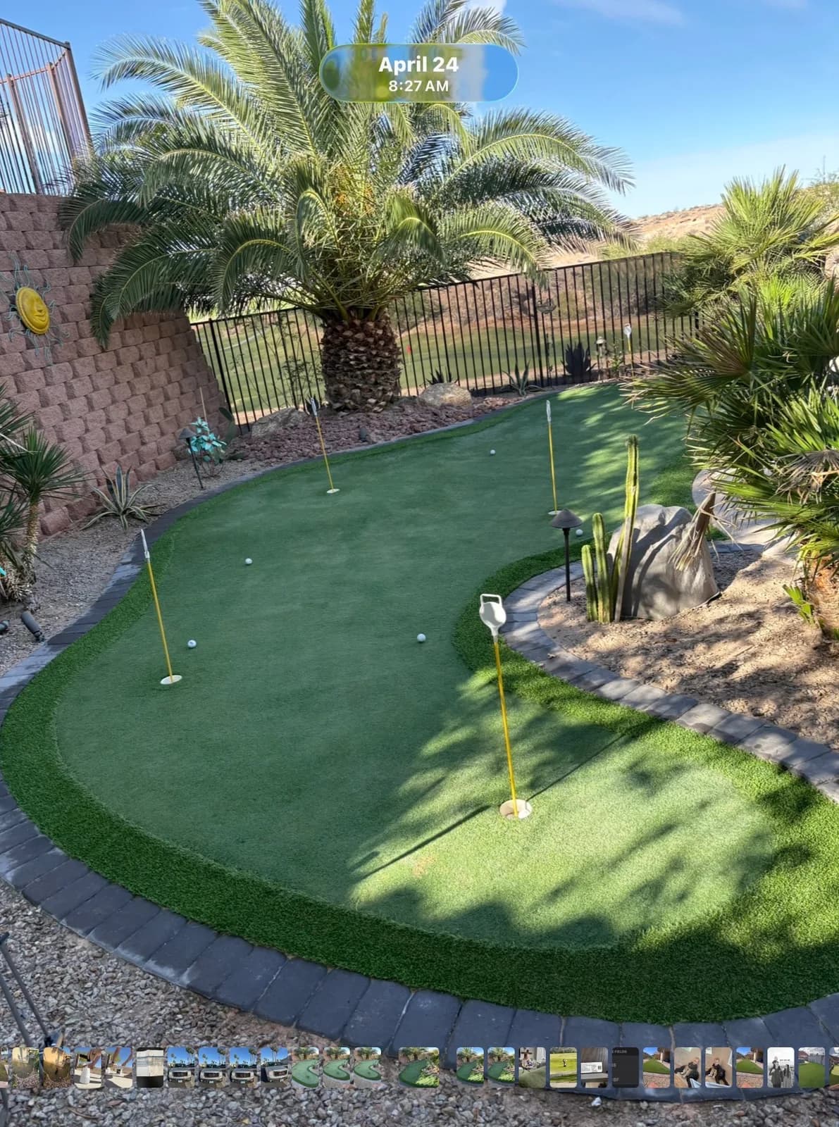 Aerial view of a clean backyard artificial turf putting green with four holes and yellow flag sticks, surrounded by desert landscaping, palm trees, and a block retaining wall in Mesquite, NV.