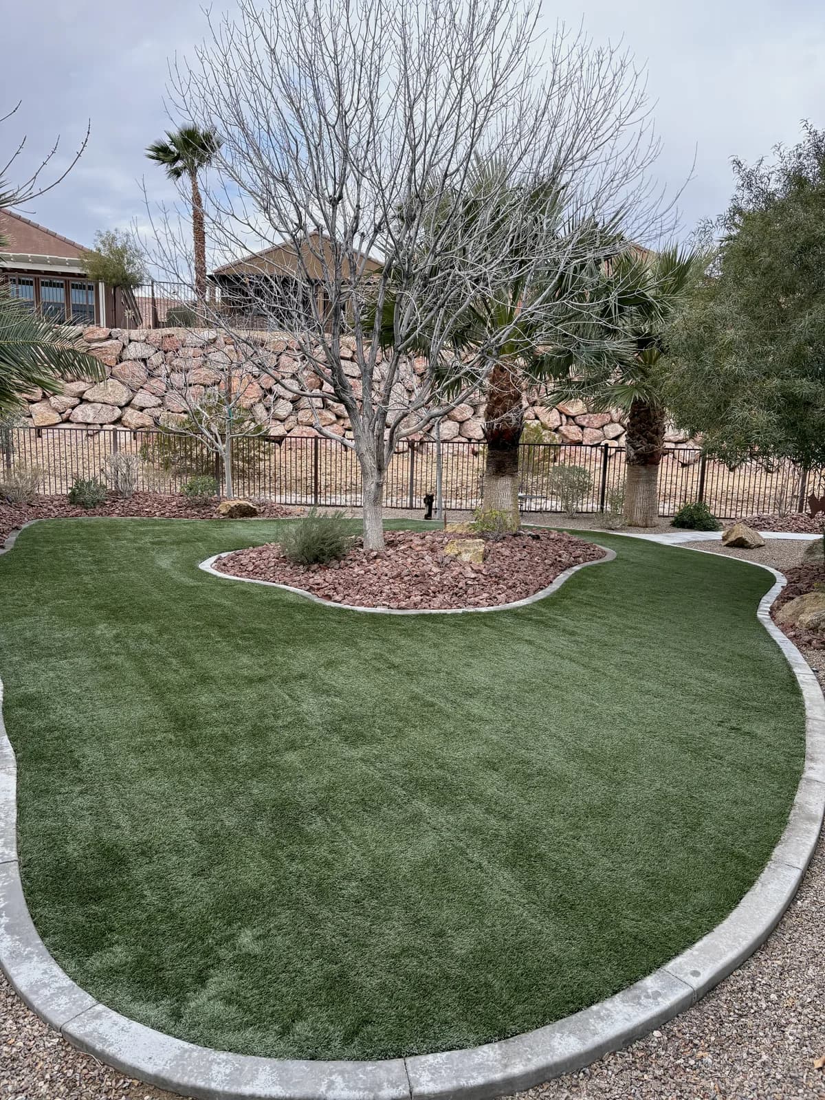 Restored artificial turf lawn curving around a desert landscaping bed with palm trees and a stone retaining wall